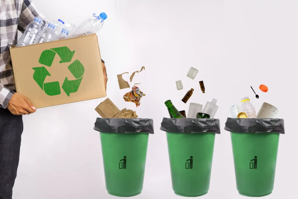 A man holding a cardboard box filled with recycled materials alongside three recycle bins.
